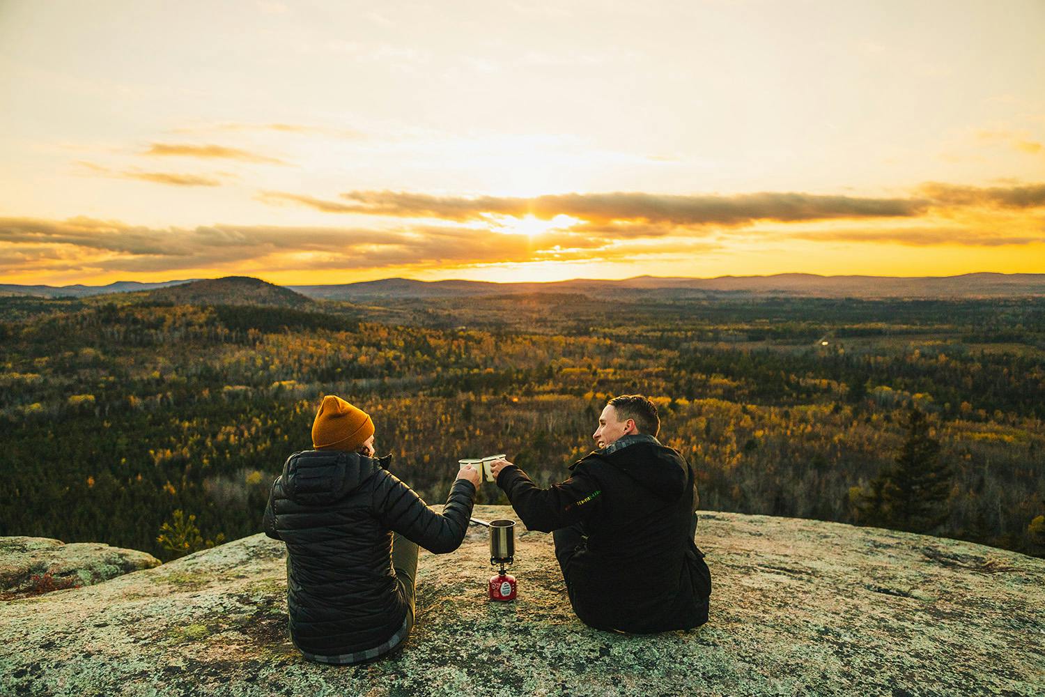 Two people sitting on a mountain in front of a sunrise with a cup of tee or coffee in their hands